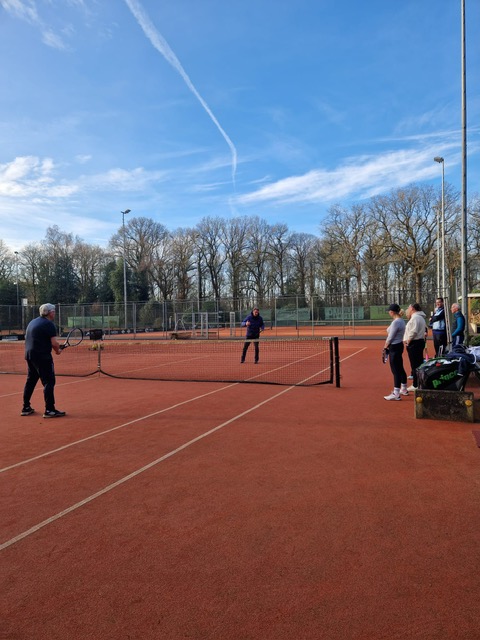Tennissers spelen samen op een gravelbaan, met de groep langs de baan